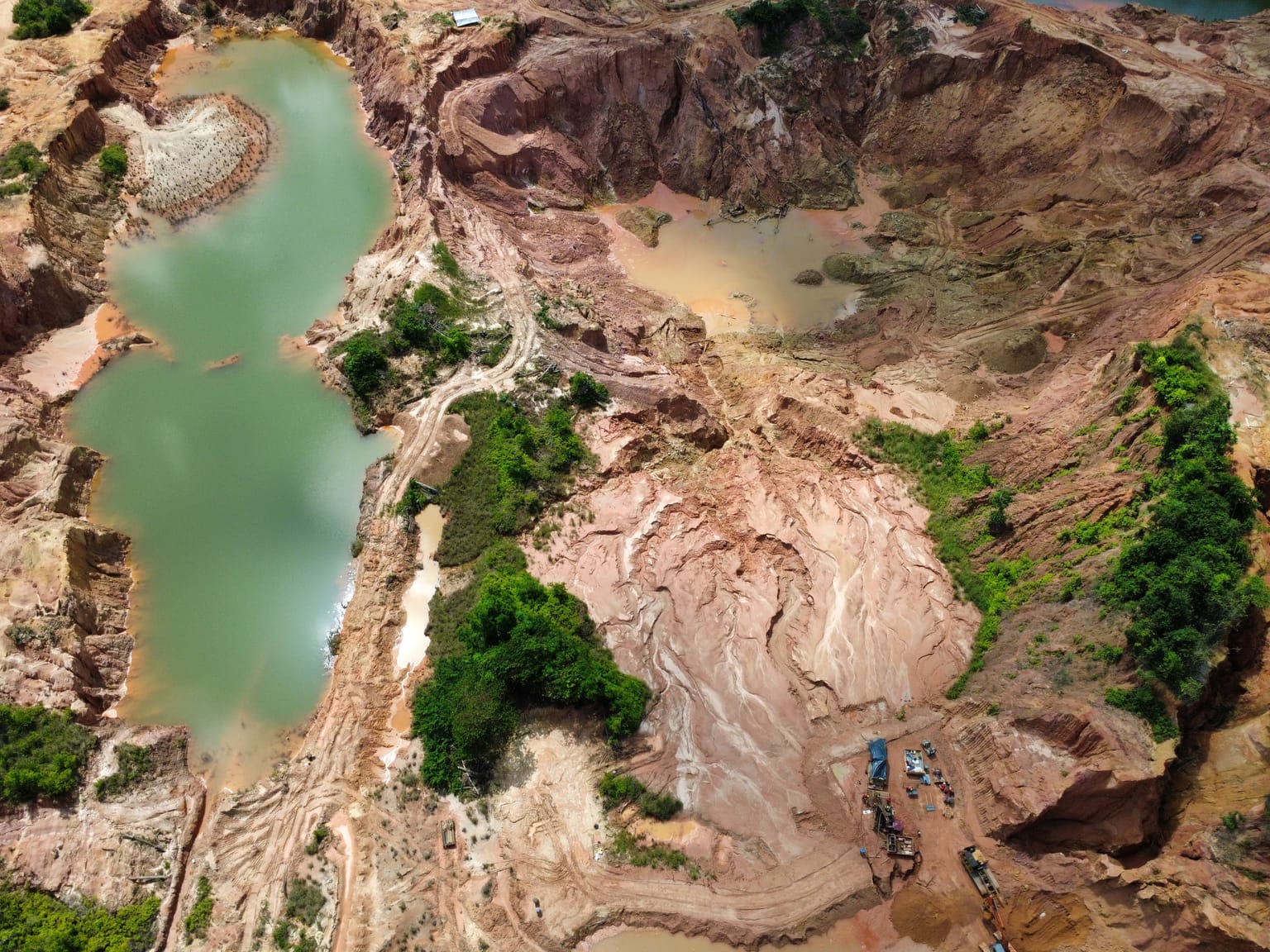 Top-down drone view of an emerald pond ringed by red laterite in a worked artisanal pit, Sela Creek