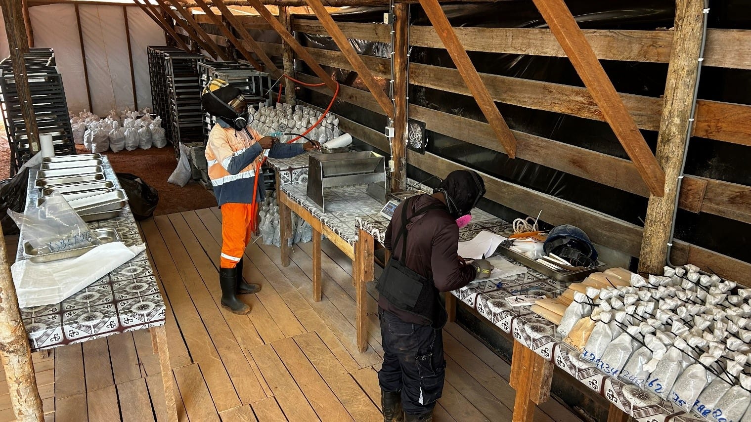 Two workers inside the Sela Creek sample preparation laboratory, a timber-framed building with bagged samples, drying trays and a crusher on workbenches.